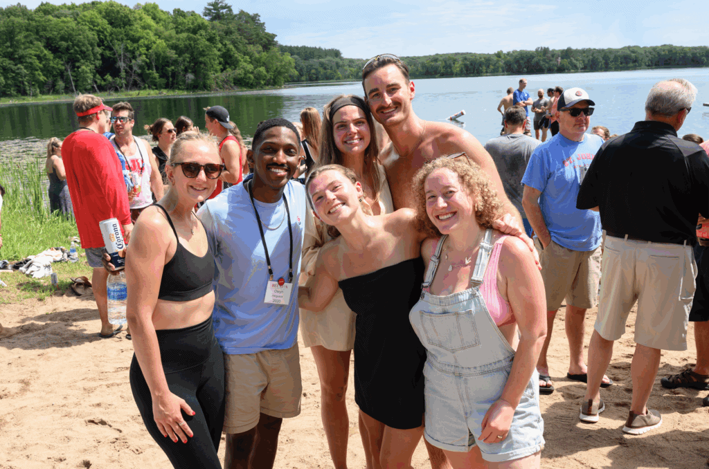 A group of six smiling young adults pose together on a sandy lakeshore, with trees and water in the background. Other people are mingling in the background, enjoying a sunny day outdoors.